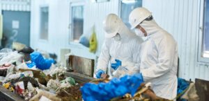 Portrait of two workers wearing biohazard suits and hardhats working at waste processing plant sorting recyclable materials on conveyor belt