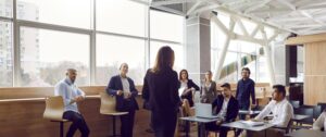 Various businessmen listen to female business leader during corporate meeting or training lecture. Rear view of woman in business attire speaking to people in modern business center. Panoramic view.