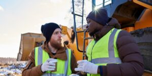 Portrait of two workers, one African-American, drinking coffee and chatting next to heavy industrial truck on worksite