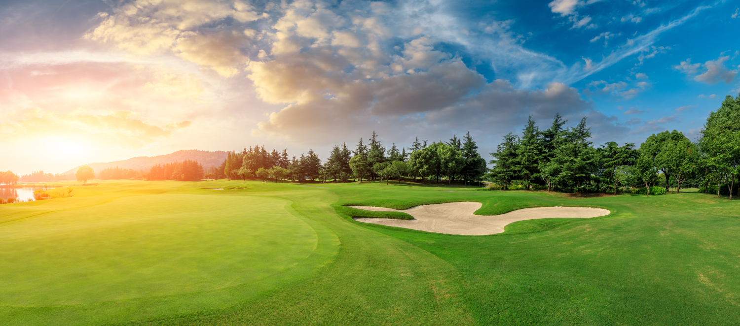 A golf course with a beautiful sunrise and well maintained green. The sun is a bright orange and casts is rays across the course. 