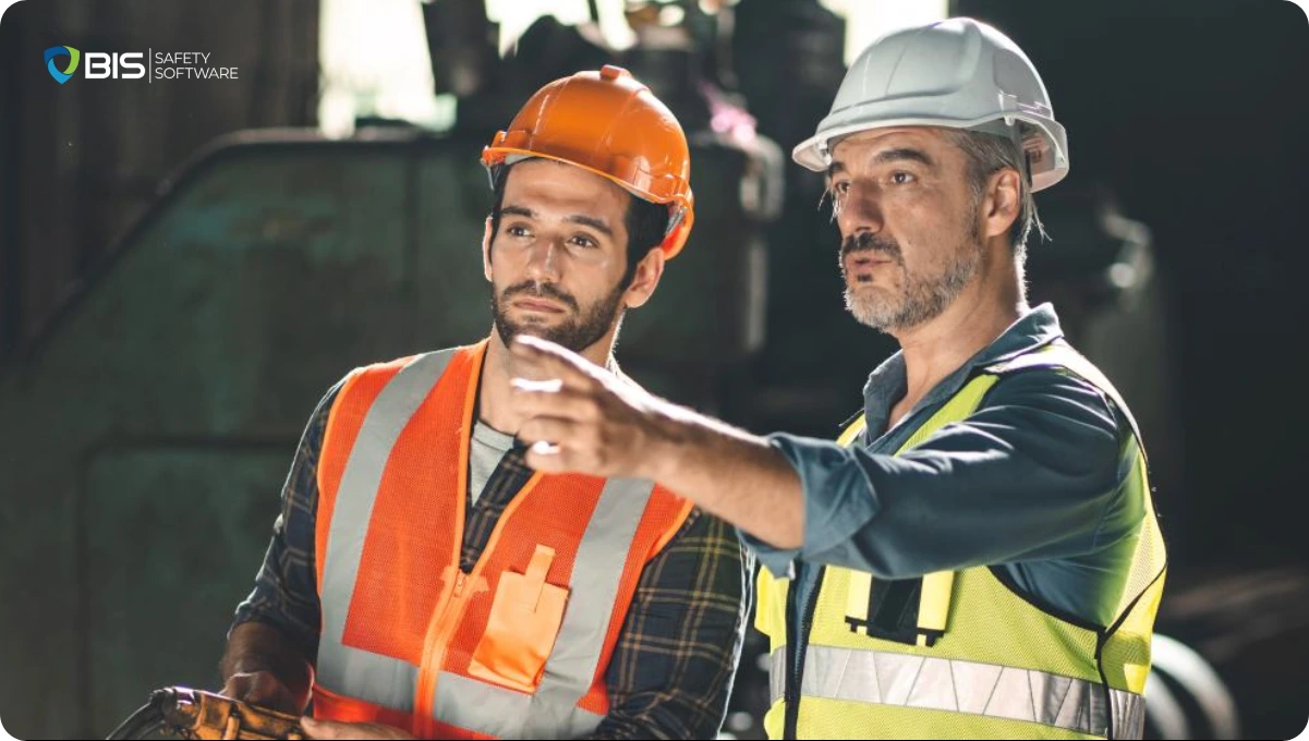 Two male industrial workers in hard hats and safety vests discuss operations inside a factory, with one pointing and giving instructions.