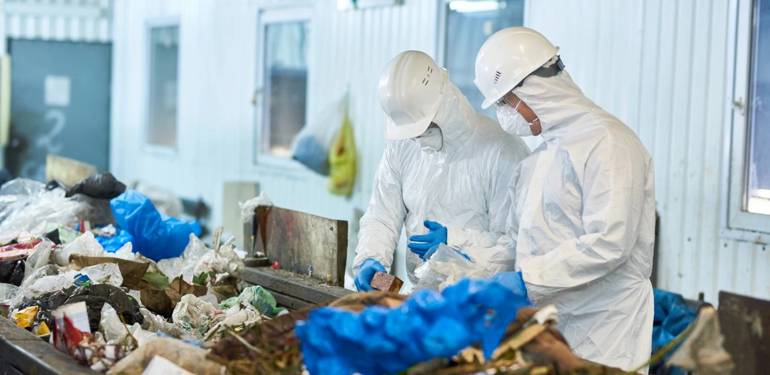 - BIS Safety Software Canada Portrait of two workers wearing biohazard suits and hardhats working at waste processing plant sorting recyclable materials on conveyor belt